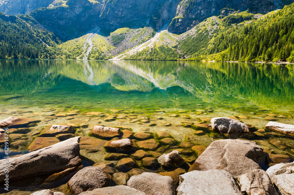 Zdjęcie przedstawia malownicze Morskie Oko w Tatrach. Krystalicznie czysta woda, skaliste brzegi, górskie szczyty w tle i zieleń lasów.