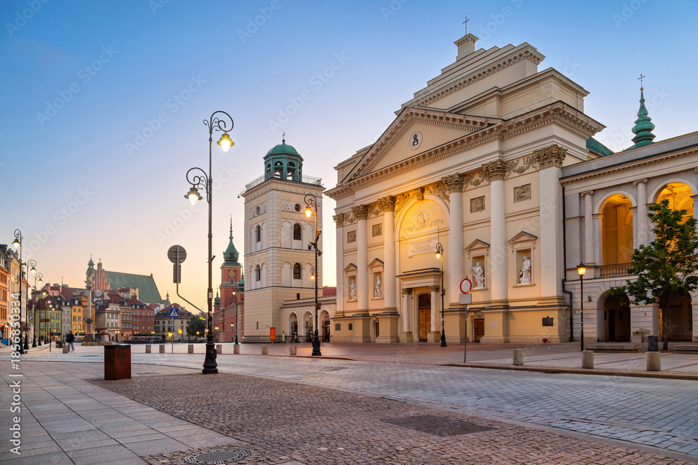 Wieczorna fotografia klasycystycznej fasady kościoła z dzwonnicą, brukowany plac, błękitne niebo i złote światła latarni - obraz drukowany na płótnie