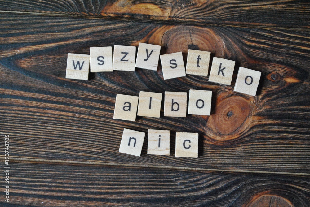 Napis 'WSZYSTKO ALBO NIC' ułożony z jasnych drewnianych płytek na ciemnym, teksturowanym drewnianym tle