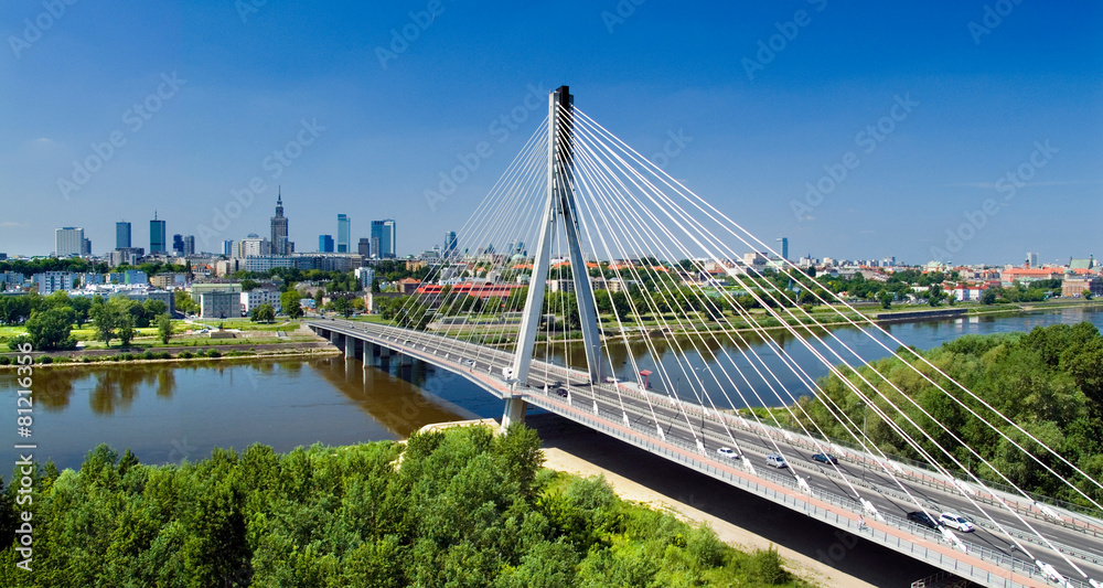 Fotografia mostu wantowego nad Wisłą z pylonami i wantami, w tle panorama Warszawy z Pałacem Kultury, błękitne niebo - obraz drukowany na płótnie